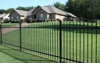 A black metal fence surrounds a large, well-kept lawn in front of a single-story brick house with a gabled roof and trimmed bushes.