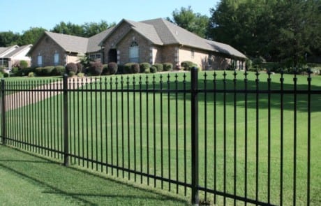 A black metal fence surrounds a large, well-kept lawn in front of a single-story brick house with a gabled roof and trimmed bushes.