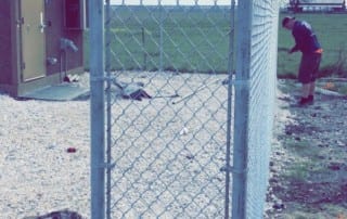 A chain-link fence section stands alone near a gravel area, with a small building to the left and a person working in the background on a grassy field.