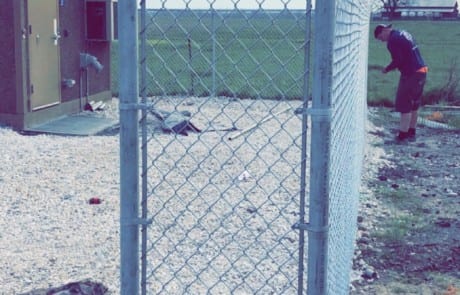 A chain-link fence section stands alone near a gravel area, with a small building to the left and a person working in the background on a grassy field.