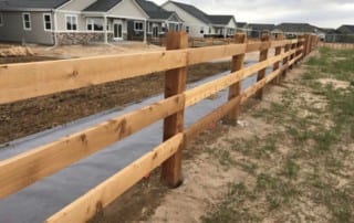 A wooden split-rail fence runs alongside a paved path in front of several suburban houses under a cloudy sky.