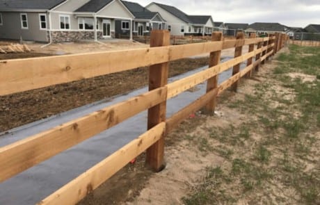 A wooden split-rail fence runs alongside a paved path in front of several suburban houses under a cloudy sky.