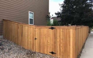 A wooden privacy fence with a gate encloses the side yard of a house, running alongside a gravel area and a concrete walkway.