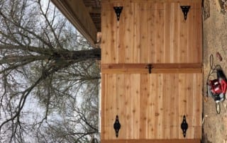 A large wooden double gate with black metal hinges and latch is installed beside a brick wall; tools are on the ground nearby.