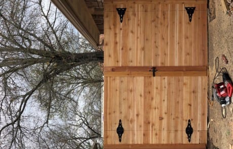 A large wooden double gate with black metal hinges and latch is installed beside a brick wall; tools are on the ground nearby.