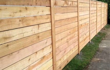 A long wooden fence made of horizontal planks runs alongside a strip of grass and a narrow concrete path.