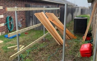 Wooden fence boards leaning on a partially constructed metal and wood frame in a narrow backyard space between two houses on a cloudy day.