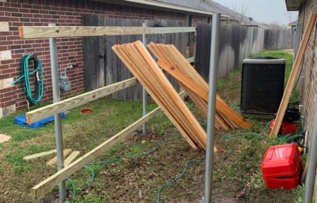 Wooden fence boards leaning on a partially constructed metal and wood frame in a narrow backyard space between two houses on a cloudy day.
