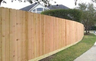 A tall, new wooden fence runs alongside a sidewalk in a suburban neighborhood with grass and trees.