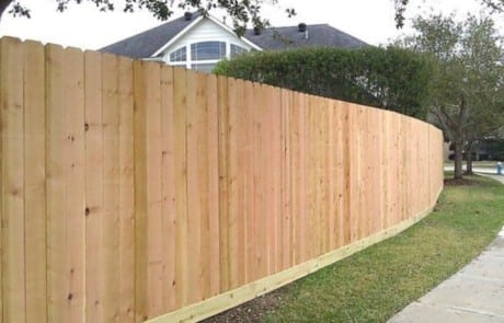 A tall, new wooden fence runs alongside a sidewalk in a suburban neighborhood with grass and trees.