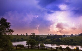 Lightning bolts strike in the distance over a rural landscape with rolling hills, trees, a river, and a wooden fence in the foreground at twilight.