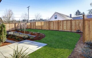 Backyard with a wooden privacy fence, green lawn, landscaped garden beds, small trees, and a stone path leading from a patio area.