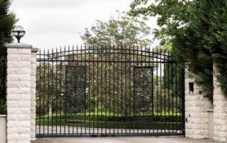 A closed black iron gate with pointed tips is set between stone pillars, surrounded by trees and greenery.