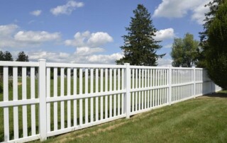 A white wooden picket fence runs along a grassy lawn with trees and a partly cloudy sky in the background.