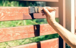 A person paints a wooden fence with a brush, applying a reddish-brown stain in sunlight.