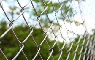 Close-up view of a metal chain-link fence with a blurred background of green trees and a partly cloudy sky.