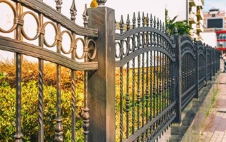 Black metal fence with decorative circular and pointed elements runs along a sidewalk next to a garden with shrubs and a building in the background.