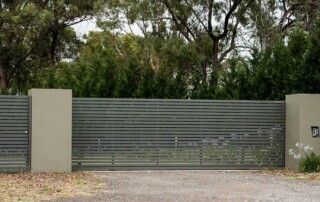 A large, horizontal metal sliding gate with matching pedestrian gate and mailbox, set in a beige wall, with tall green trees in the background.