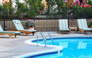 Four striped lounge chairs sit by a circular outdoor swimming pool, with potted flowers and a black metal fence in the background.