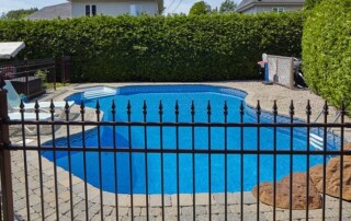 A private backyard swimming pool is surrounded by a black metal fence with trees and houses in the background.