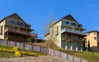 Three modern, two-story houses on a sloped hill with wooden fences and green shrubbery under a clear blue sky.