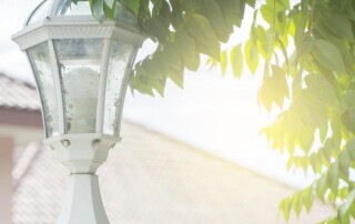 A white outdoor lantern with a glass cover stands next to green tree branches, with sunlight shining through the leaves in the background.
