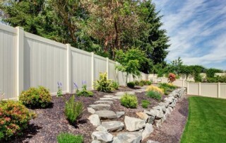 Well-maintained backyard with stone pathway, mulched garden beds, small shrubs, and a tall white privacy fence bordering the lawn.