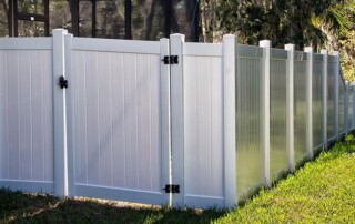 White vinyl privacy fence with gate panels runs alongside a building, set on grass with trees in the background.