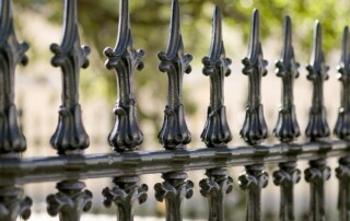 Close-up view of a black metal fence with decorative pointed finials, set outdoors with blurred greenery in the background.