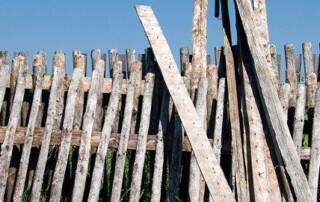 A weathered wooden fence with uneven slats stands outdoors, with several additional wooden boards leaning against it under a clear blue sky.
