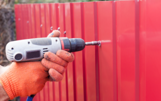 A person wearing orange gloves uses a power drill to fasten screws into a red metal sheet fence.