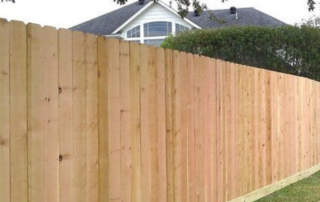 A tall wooden privacy fence runs along a yard, with a house and trees visible in the background.
