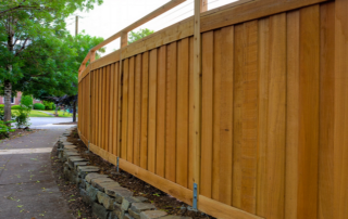 A tall wooden privacy fence runs alongside a sidewalk bordered by a stone retaining wall, with trees and houses visible in the background.