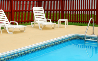 A swimming pool with metal steps, surrounded by a concrete patio, with three white lounge chairs and a small table near a red wooden fence.