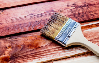 A paintbrush with a wooden handle applies stain to wooden planks, partially coloring the surface a rich brown shade.