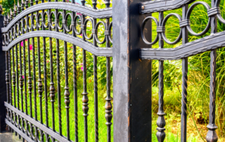 Black wrought iron fence with decorative circular and vertical patterns, bordered by green plants and grass in the background.