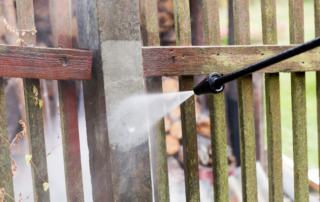 A person uses a pressure washer to clean a dirty wooden fence, revealing a lighter, cleaner section compared to the surrounding wood.