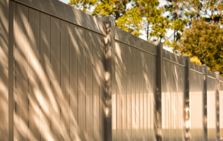 A tall, beige vinyl fence with vertical panels casts shadows from nearby trees on a sunny day. Trees with green foliage are visible in the background.