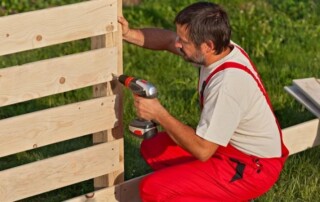 A man in red overalls uses a cordless drill to assemble a wooden fence outdoors on grass.