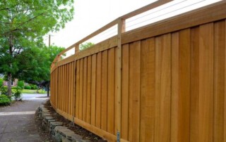 Tall wooden privacy fence with vertical planks runs alongside a sidewalk, bordered by a stone edging and trees.