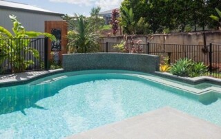 Outdoor swimming pool with clear water, surrounded by a black metal fence, tropical plants, and a tiled poolside area on a sunny day.