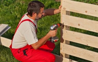 A man in red overalls uses a cordless drill to assemble a wooden fence outdoors on a grassy area.