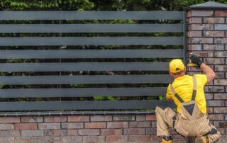 A worker in yellow clothing installs or repairs a modern horizontal metal fence attached to a brick pillar outdoors.