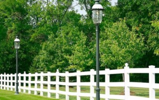 White wooden fence runs alongside a row of vintage-style black street lamps with green trees and grass in the background.