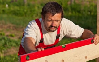 Man in red overalls uses a spirit level to check the alignment of a wooden plank outdoors, with greenery in the background.