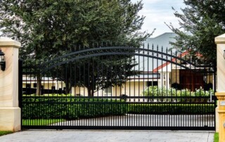 A black wrought iron gate with stone pillars and lantern lights stands closed at the entrance of a property with trees and a house in the background.