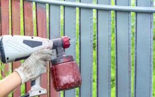 A person wearing a work glove uses a paint sprayer to paint a metal fence gray.