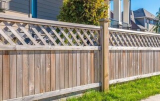 A wooden fence with lattice panels on top runs alongside a green grassy area near a sidewalk in a residential neighborhood.