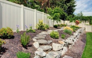 A landscaped backyard with a stone pathway, various shrubs, mulch, and a white vinyl privacy fence under a partly cloudy sky.