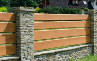 A modern fence made of horizontal wooden planks and stone pillars encloses a grassy yard with a house and greenery in the background.